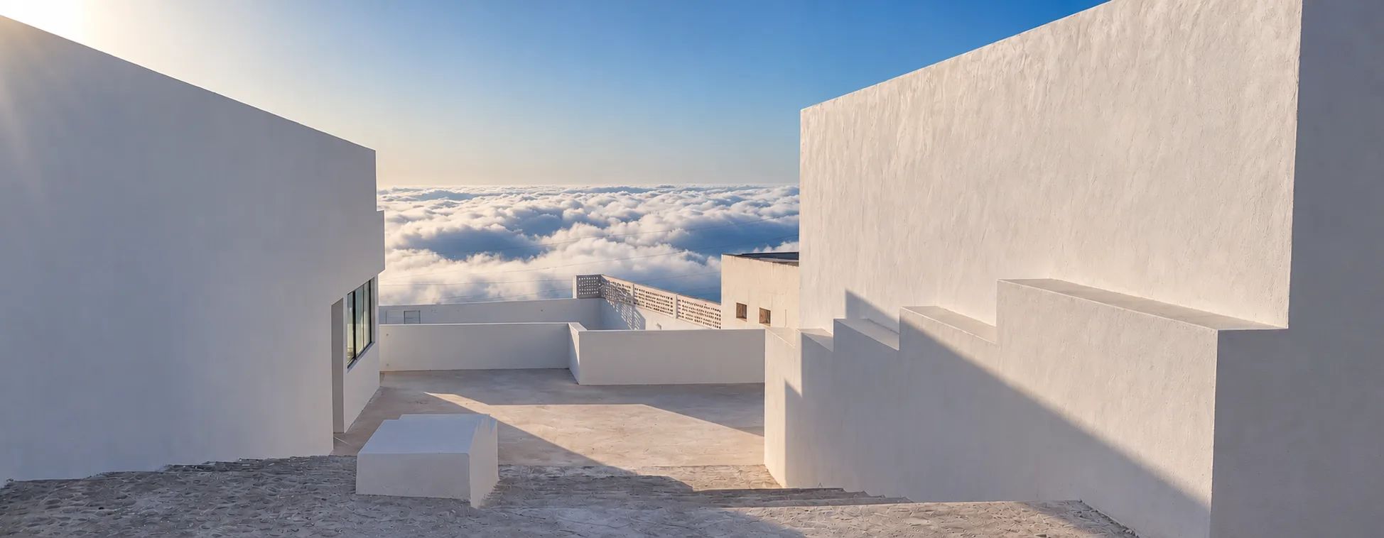 Weiße Mauern und Terrasse über einem Wolkenmeer, darüber blauer Himmel